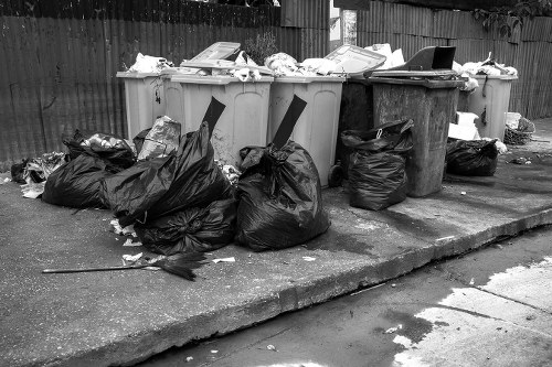 A row of five large waste bins positioned on a paved driveway, with two yellow, one grey, and one black container, all filled with discarded rubbish. Several black trash bags are piled on the ground in front of the bins, some partially torn and spilling contents. The bins and bags contain a mix of paper, cardboard, plastic packaging, and miscellaneous household waste. The scene is outdoors, with a wooden fence visible in the background. The overall setting suggests a typical rubbish collection point in an urban residential area, consistent with waste management services provided by Garden Clearance Wanstead in the Wanstead region, within the E11 postcode area. Natural daylight illuminates the scene, highlighting the textures of the waste materials, with some clutter hinting at recent rubbish clearance activity. This image exemplifies the kind of waste that Garden Clearance Wanstead manages for local residents, removing household and garden refuse efficiently and responsibly.