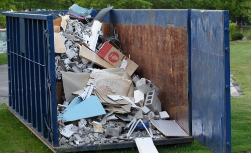 A large industrial metal skip painted deep blue with rusted interior walls, positioned outdoors on a grassy area near a paved surface. The skip is filled with a mixture of construction and renovation debris, including white and grey plasterboard, broken tiles, fragments of concrete, and assorted discarded materials. Some waste items, such as a corrugated cardboard box and broken ceramic pieces, are visible amongst the pile. The debris extends above the height of the skip’s edges, indicating it is nearly full. In the background, blurred greenery and trees suggest a suburban or park-like setting, typical of the Wanstead area. The image portrays a scene consistent with professional rubbish collection or garden clearance services, such as those provided by Garden Clearance Wanstead, emphasizing the removal of building waste and general rubbish. Natural daylight illuminates the scene, highlighting the textures of the debris and the weathered surface of the skip, aligning with waste management work typical in local residential or garden environments in East London.