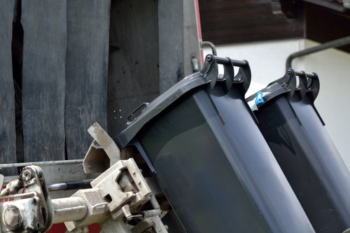 Close-up of two dark grey wheelie bins with black lids, positioned on a metal frame and attached to a vehicle or trolley, with part of a grey wall or wooden fencing visible in the background. The bins are made of durable plastic with a smooth finish and are placed outdoors, likely for rubbish collection or waste disposal in Wanstead. The environment suggests a residential or commercial setting with a practical arrangement for managing waste. The image emphasizes the bins’ sturdy construction and functional design, typical of waste removal services offered by Garden Clearance Wanstead, who provide professional rubbish removal in the local area.