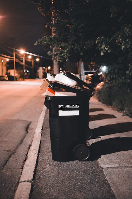 A waste management worker wearing a high-visibility yellow and red uniform operates a large, red rubbish collection vehicle on a street. The back of the vehicle is open, revealing the compaction mecha
