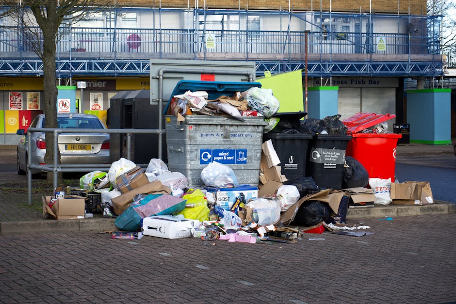 A close-up view of a pile of mixed waste materials on the ground, consisting of crumpled paper, plastic bottles, cardboard packaging, and torn paper bags. The plastic bottles are transparent with labe